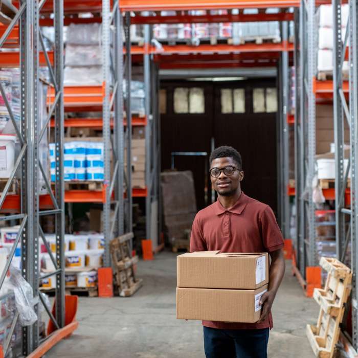 Positive young African-American storeroom worker in glasses walking with boxes over warehouse and carrying order to customer